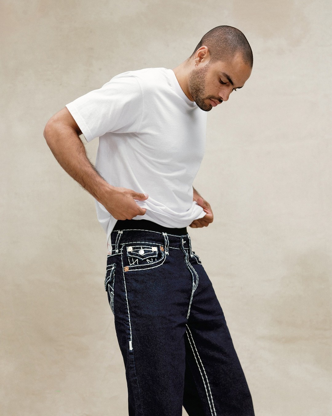 Male model looking downwards and lifting his shirt slightly to display his jeans against a tan backdrop.